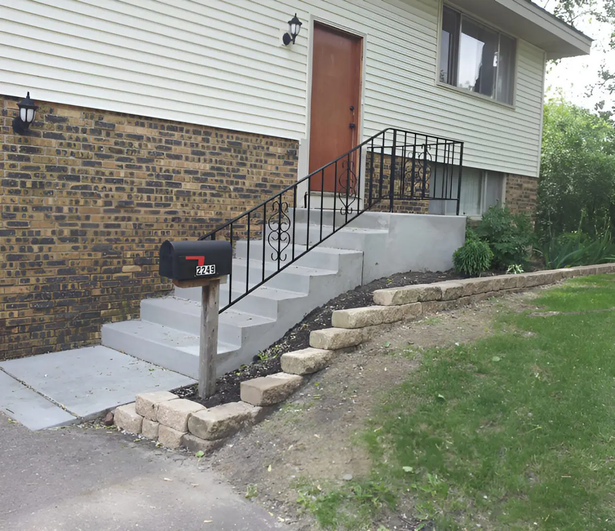 Concrete steps with metal railing leading to a wooden door of a brick house. A mailbox with the number "2245" is next to a small retaining wall and a grassy yard.